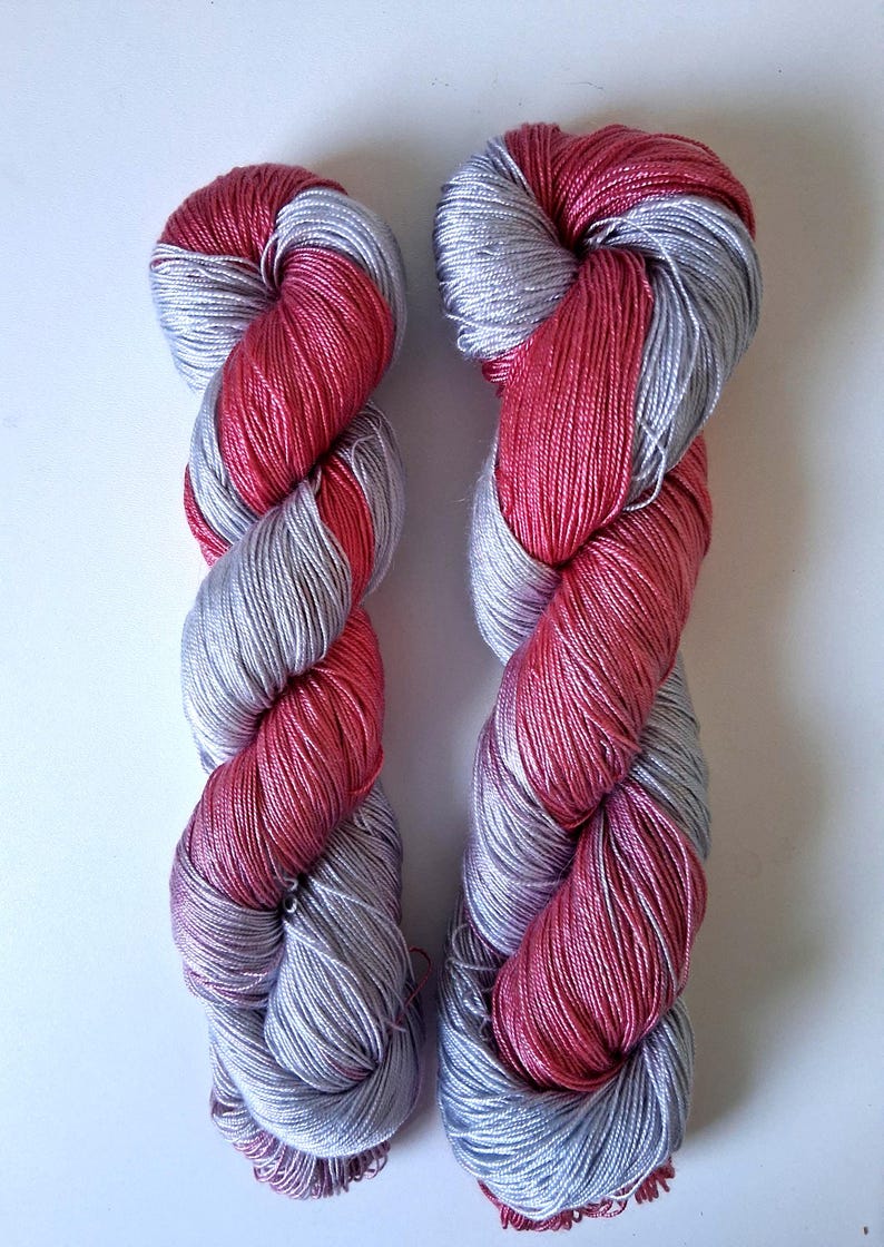 Puede incluir: Dos madejas de hilo en un patr&oacute;n retorcido. El hilo es una combinaci&oacute;n de plata y un color rosa-rojo vibrante. Las madejas se muestran sobre un fondo blanco liso, resaltando los colores y la textura del hilo.