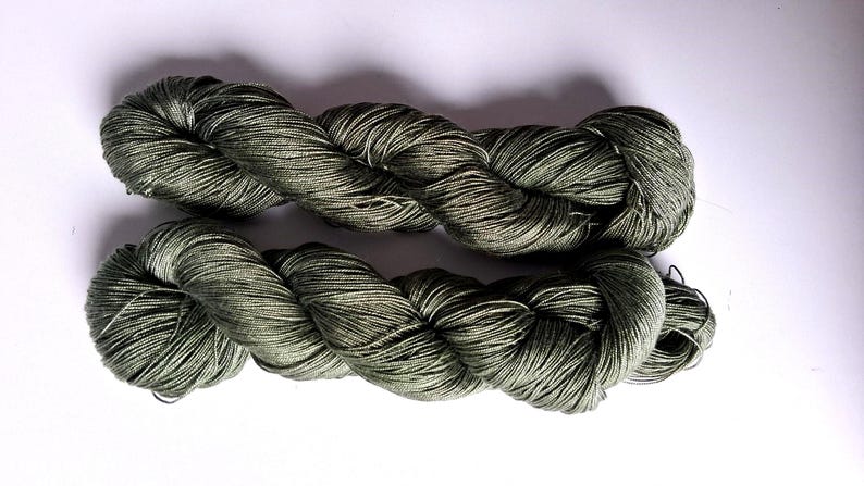 May include: Two skeins of olive green yarn are shown against a white backdrop. The yarn is tightly twisted, with a subtle sheen. The texture suggests a soft, natural fibre, suitable for knitting or crochet projects.