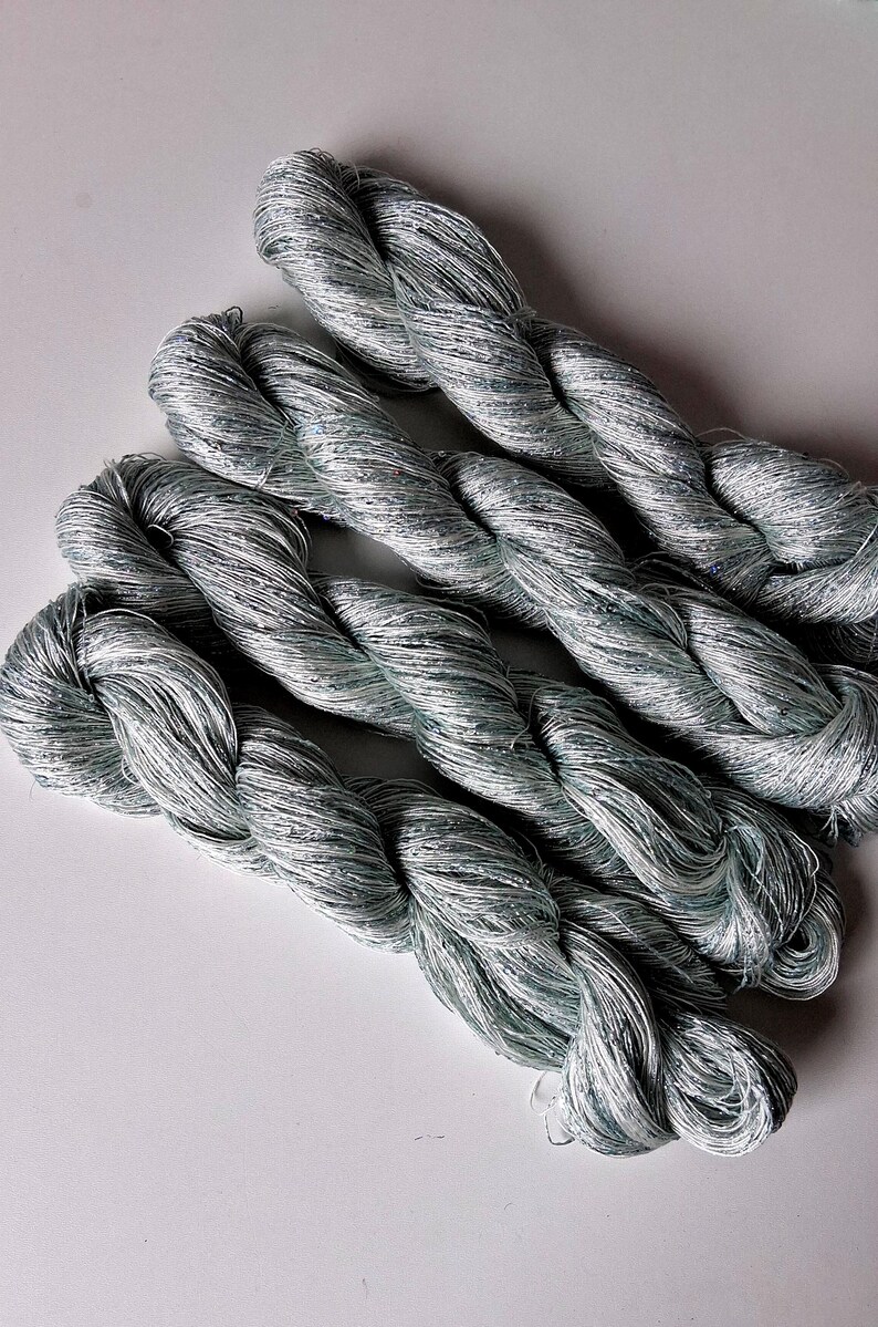 May include: Close-up of several skeins of silver-gray yarn. The yarn is tightly twisted, with a slight sheen and a subtle sparkle. The texture appears soft and pliable, suggesting it is suitable for various crafting projects.