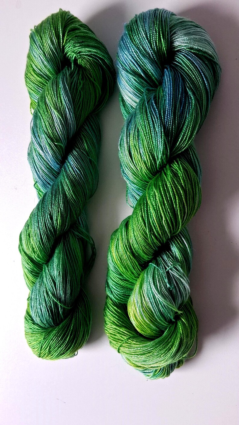 May include: Two skeins of vibrant green yarn with subtle blue and yellow highlights. The yarn is tightly twisted, creating a textured appearance. The color gradient adds depth and visual interest, perfect for knitting or crochet projects.