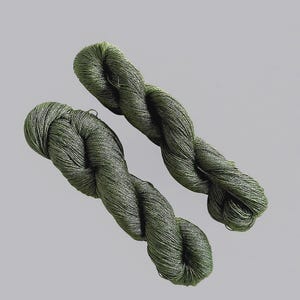 May include: Two skeins of olive green yarn are shown against a light grey backdrop. The yarn is tightly twisted, showing a textured surface. The colour is a muted, earthy green, suitable for various crafting projects. The yarn appears soft and flexible.
