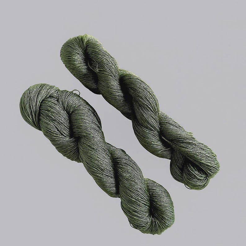 May include: Two skeins of olive green yarn are shown against a light grey backdrop. The yarn is tightly twisted, showing a textured surface. The colour is a muted, earthy green, suitable for various crafting projects. The yarn appears soft and flexible.