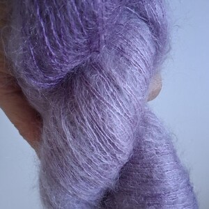 Puede incluir: Una madeja de hilo suave y difuso en un tono p&uacute;rpura vibrante. La fibra tiene un efecto halo distintivo, caracterizado por una textura fina y delicada con hebras visibles retorcidas para crear una apariencia voluminosa y aireada.