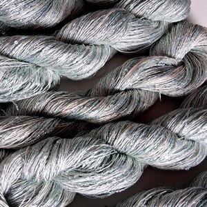May include: Close-up of several skeins of silver-gray yarn. The yarn is tightly twisted, with a slight sheen and a subtle sparkle. The yarn appears to be made of a soft, fine material, suitable for knitting or crocheting projects. The overall theme is craft supplies.