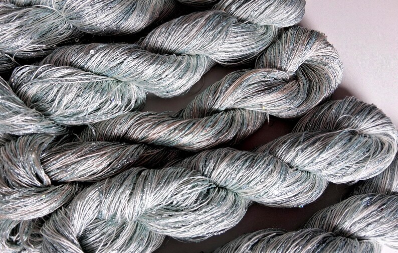 May include: Close-up of several skeins of silver-gray yarn. The yarn is tightly twisted, with a slight sheen and a subtle sparkle. The yarn appears to be made of a soft, fine material, suitable for knitting or crocheting projects. The overall theme is craft supplies.