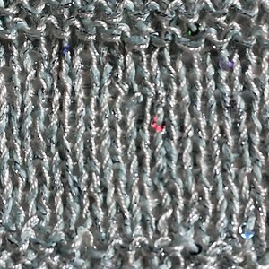 May include: Close-up of a knitted fabric in a light gray color with a subtle shimmer. The knit has a textured appearance with vertical ribbing and small, colorful sequins scattered throughout. The fabric appears soft and pliable.