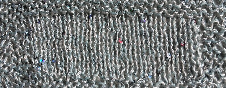 May include: Close-up of a knitted fabric in a light gray color with a subtle shimmer. The knit has a textured appearance with vertical ribbing and small, colorful sequins scattered throughout. The fabric appears soft and pliable.