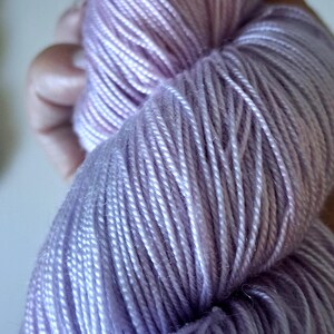 May include: Close-up of a skein of lavender yarn. The yarn is tightly wound, showing individual strands with a slight sheen. The colour transitions from a deeper purple to a lighter, almost white hue.