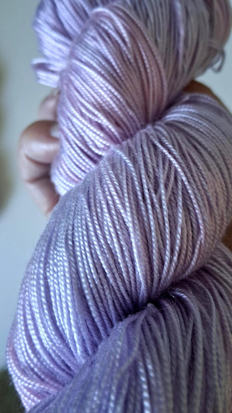 May include: Close-up of a skein of lavender yarn. The yarn is tightly wound, showing individual strands with a slight sheen. The colour transitions from a deeper purple to a lighter, almost white hue.