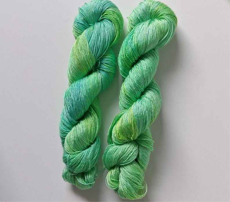 Peut inclure: Deux &eacute;cheveaux de fil teints &agrave; la main dans des tons de vert et de bleu. Le fil est torsad&eacute; en forme de corde, avec les couleurs se fondant les unes dans les autres. Le fil semble doux et d&eacute;licat, adapt&eacute; aux projets de tricot ou de crochet.