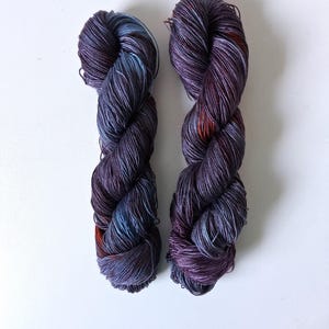May include: Two skeins of hand-dyed yarn in shades of purple, blue, and orange. The yarn is tightly wound and has a slight sheen, suggesting a silk or similar fiber. The colors blend together, creating a variegated effect. The yarn is displayed against a white background.