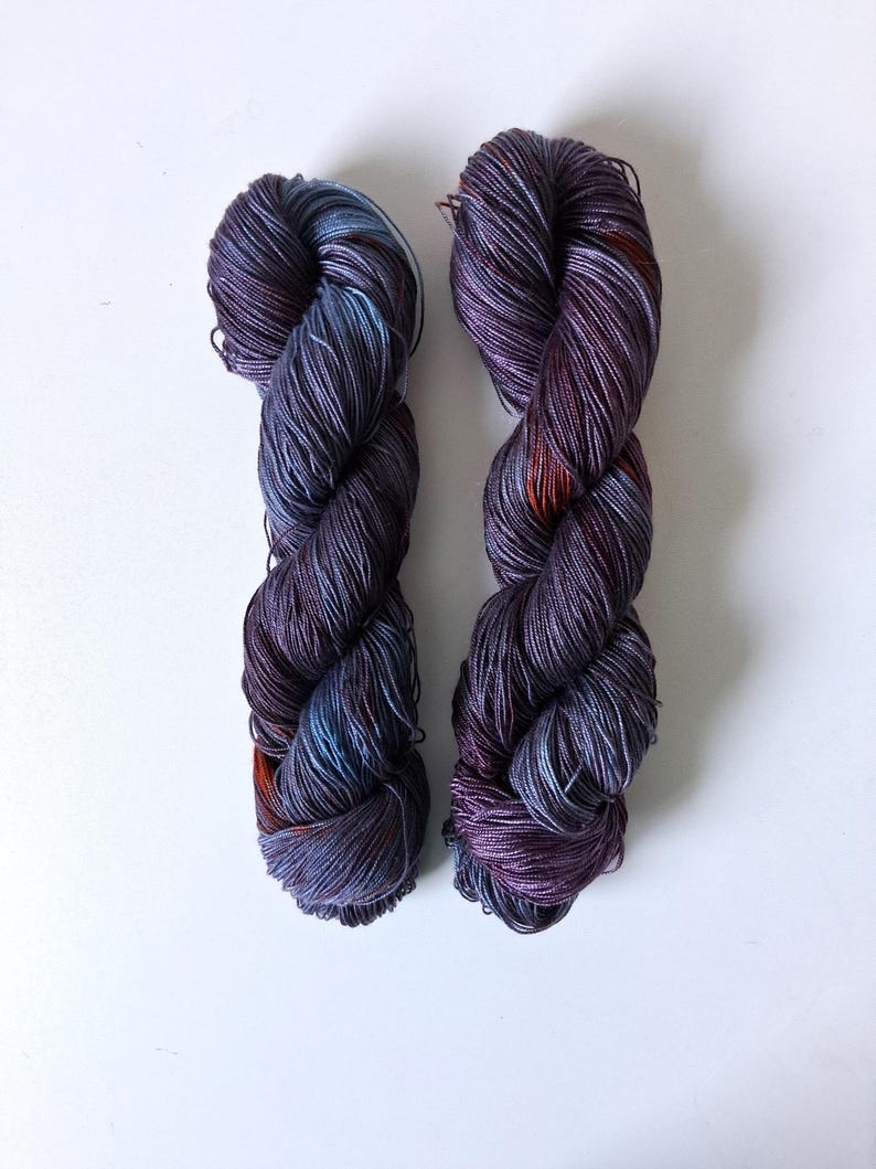 May include: Two skeins of hand-dyed yarn in shades of purple, blue, and orange. The yarn is tightly wound and has a slight sheen, suggesting a silk or similar fiber. The colors blend together, creating a variegated effect. The yarn is displayed against a white background.