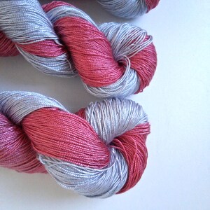 Puede incluir: Primer plano de dos madejas de hilo. El hilo es una combinaci&oacute;n de plata y un color rosa intenso. El hilo est&aacute; torcido y enrollado, mostrando las variaciones de textura y color. El fondo es una superficie blanca lisa.