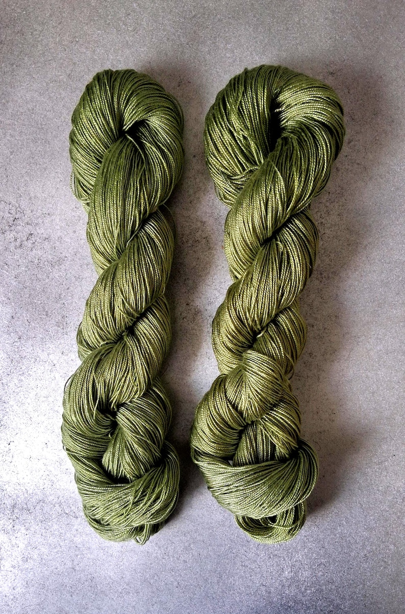 May include: Two skeins of olive green yarn are displayed against a gray background. The yarn is tightly twisted, revealing the individual strands. The color is a muted, earthy green, suggesting a natural fiber. The yarn is likely used for knitting or crocheting.