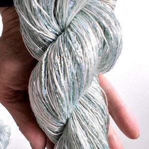 May include: A close-up of a skein of yarn in shades of light blue, white, and silver. The yarn is twisted and held in a hand, showcasing its texture and the subtle sparkle of the metallic threads. The yarn appears soft and delicate.