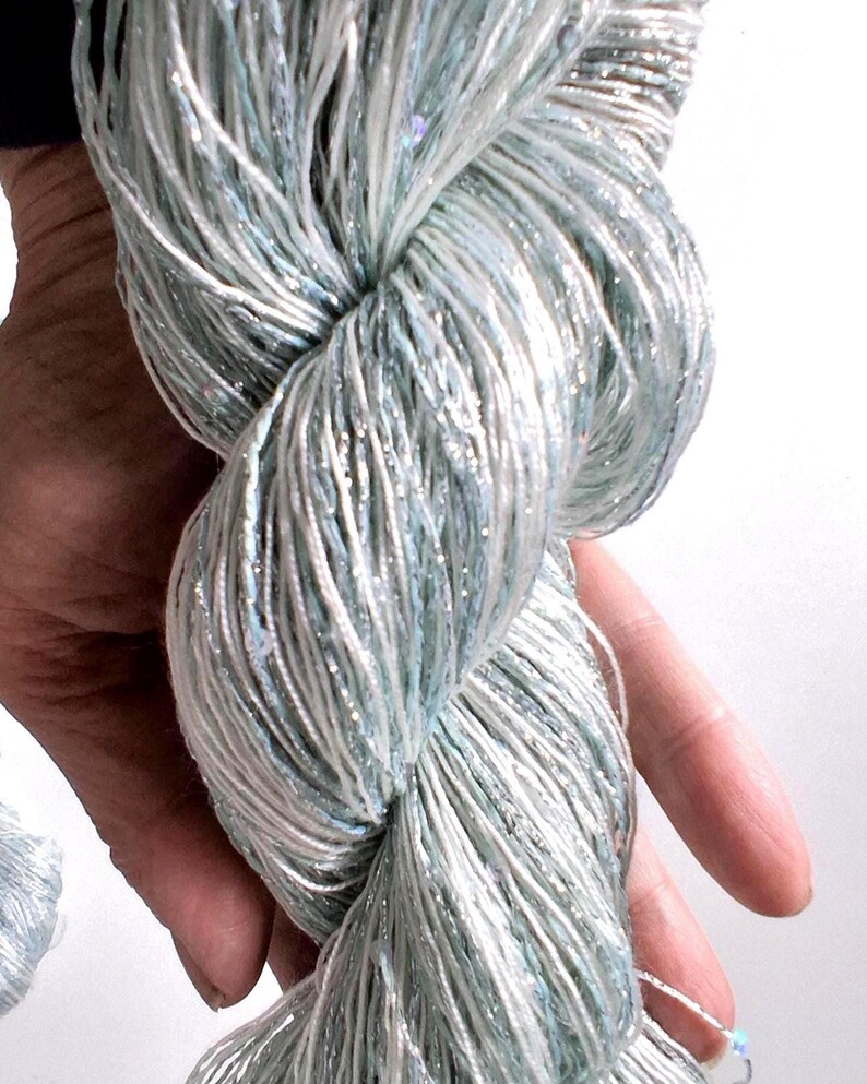 May include: A close-up of a skein of yarn in shades of light blue, white, and silver. The yarn is twisted and held in a hand, showcasing its texture and the subtle sparkle of the metallic threads. The yarn appears soft and delicate.