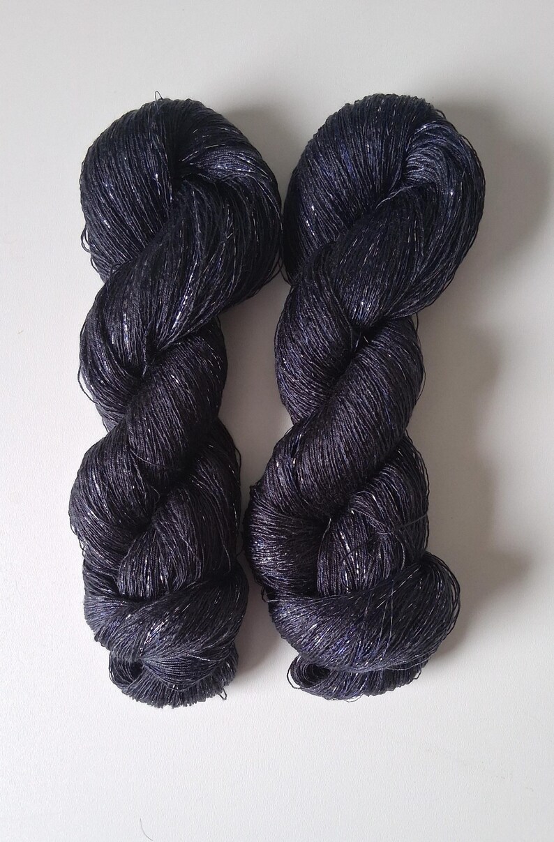 May include: Two skeins of dark navy blue yarn with a subtle metallic silver thread woven throughout. The yarn is twisted into a rope-like shape, and the texture of the yarn is visible. The background is plain white.