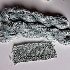 May include: Two skeins of silver-blue yarn with a slight sheen, next to a small knitted swatch. The yarn is tightly twisted, and the knitted piece shows the texture of the yarn. The color is a mix of silver and blue.