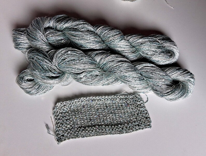 May include: Two skeins of silver-blue yarn with a slight sheen, next to a small knitted swatch. The yarn is tightly twisted, and the knitted piece shows the texture of the yarn. The color is a mix of silver and blue.