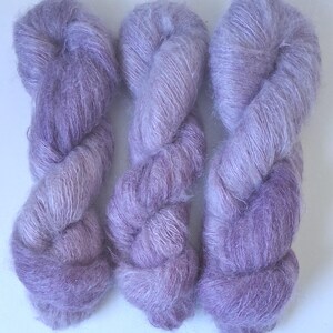 Puede incluir: Tres madejas de hilo de color morado claro y aspecto esponjoso dispuestas verticalmente. El hilo tiene una textura suave y cepillada con un efecto halo, mostrando variaciones en tonos lavanda y lila a lo largo de las hebras retorcidas.