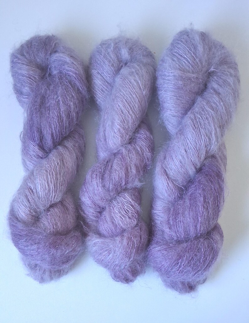 Puede incluir: Tres madejas de hilo de color morado claro y aspecto esponjoso dispuestas verticalmente. El hilo tiene una textura suave y cepillada con un efecto halo, mostrando variaciones en tonos lavanda y lila a lo largo de las hebras retorcidas.