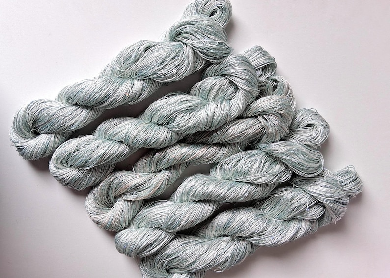 May include: Several skeins of light blue and silver yarn are arranged on a white surface. The yarn appears to be a soft, textured material, possibly silk or a similar fiber. The skeins are twisted and coiled.
