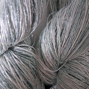May include: Close-up of several skeins of yarn. The yarn is a mix of light blue, white, and silver threads, creating a shimmering effect. The yarn appears soft and is tightly wound, suggesting it's ready for knitting or crocheting projects.