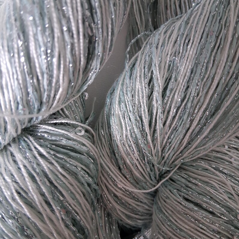 May include: Close-up of several skeins of yarn. The yarn is a mix of light blue, white, and silver threads, creating a shimmering effect. The yarn appears soft and is tightly wound, suggesting it's ready for knitting or crocheting projects.