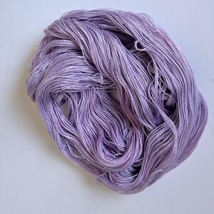 May include: A skein of lavender-coloured thread, tightly wound. The thread appears to be silk or a similar fine material, with a slight sheen. The threads are arranged in a loose, irregular shape, suitable for crafts like embroidery or sewing.