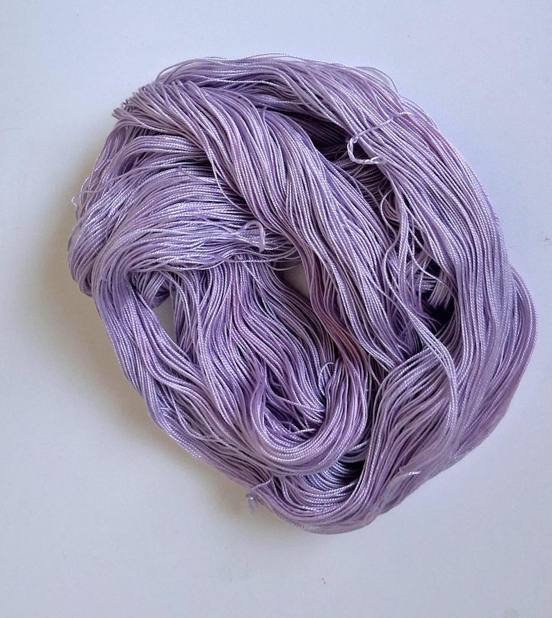 May include: A skein of lavender-coloured thread, tightly wound. The thread appears to be silk or a similar fine material, with a slight sheen. The threads are arranged in a loose, irregular shape, suitable for crafts like embroidery or sewing.