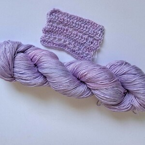 May include: A skein of lavender yarn with pink and blue hues, alongside a small crocheted swatch. The yarn is tightly wound and looks soft. The crocheted piece has an open, lacy design.