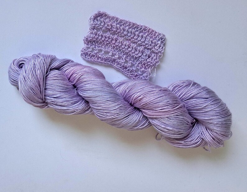 May include: A skein of lavender yarn with pink and blue hues, alongside a small crocheted swatch. The yarn is tightly wound and looks soft. The crocheted piece has an open, lacy design.