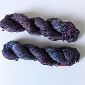May include: Two skeins of hand-dyed yarn in shades of purple, blue, and orange. The yarn is twisted into a rope-like shape, with visible texture and color variations. The yarn is likely used for knitting or crocheting projects.