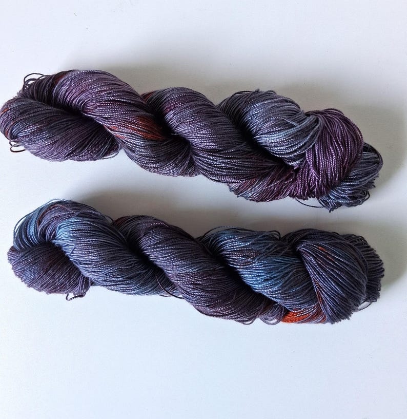 May include: Two skeins of hand-dyed yarn in shades of purple, blue, and orange. The yarn is twisted into a rope-like shape, with visible texture and color variations. The yarn is likely used for knitting or crocheting projects.
