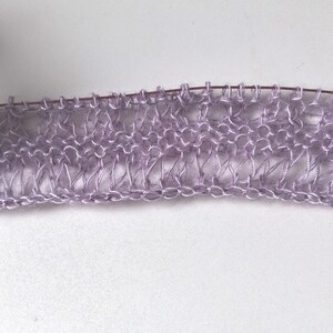 Puede incluir: Una pieza de tejido de punto color violeta claro con un patr&oacute;n de encaje calado de puntos alargados, sostenida en una aguja de tejer met&aacute;lica fina. El hilo tiene un brillo sutil y presenta una textura delicada, aireada y semitransparente.