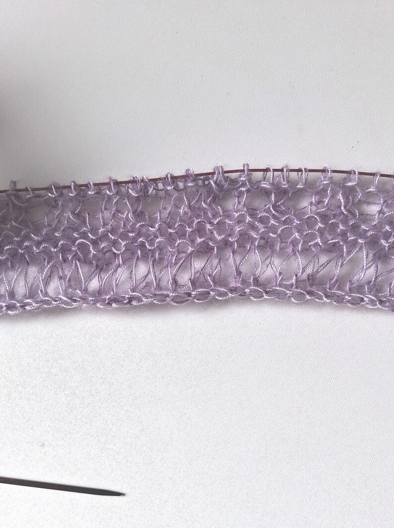 Puede incluir: Una pieza de tejido de punto color violeta claro con un patr&oacute;n de encaje calado de puntos alargados, sostenida en una aguja de tejer met&aacute;lica fina. El hilo tiene un brillo sutil y presenta una textura delicada, aireada y semitransparente.