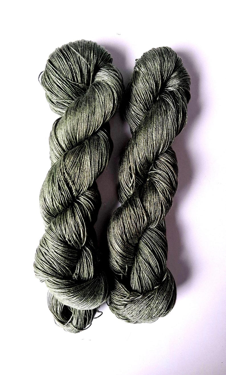 May include: Two skeins of olive green yarn are shown against a white backdrop. The yarn is tightly twisted, with a subtle sheen. The colour is a muted, earthy green, suitable for knitting or crochet projects.