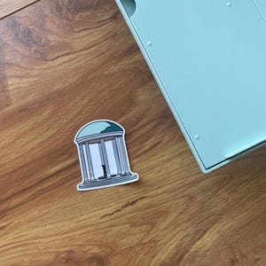 May include: A sticker featuring a white and gray illustration of a building with columns and a blue roof. The sticker is on a wooden surface, near a light blue object.