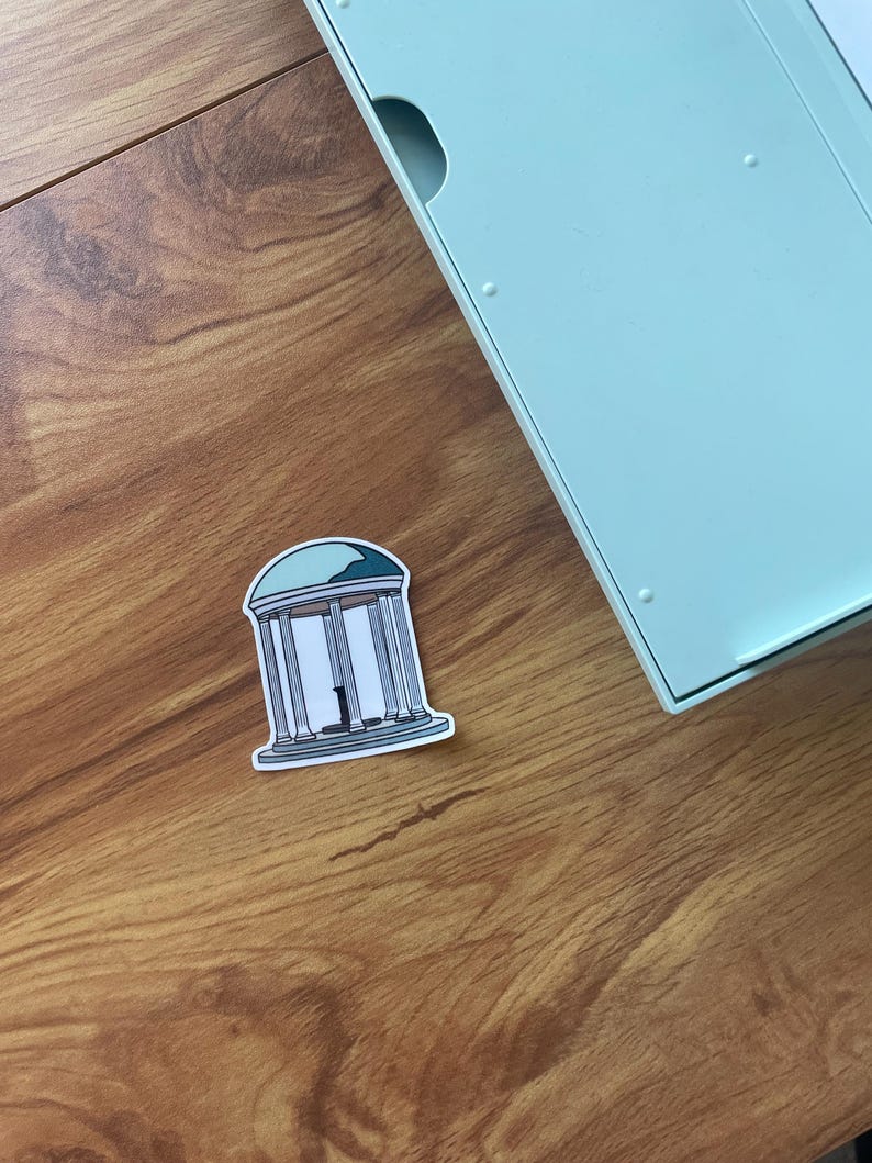 May include: A sticker featuring a white and gray illustration of a building with columns and a blue roof. The sticker is on a wooden surface, near a light blue object.
