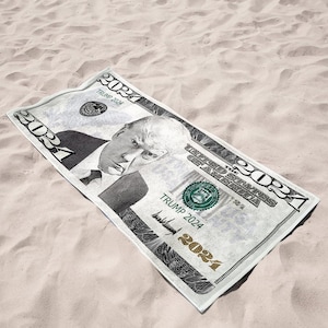 May include: A rectangular beach towel resembling a U.S. dollar bill, featuring a portrait and the text "TRUMP 2024". The towel is primarily white with black and green accents, lying on a sandy surface.
