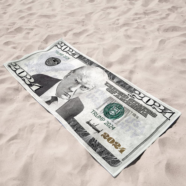 May include: A rectangular beach towel resembling a U.S. dollar bill, featuring a portrait and the text "TRUMP 2024". The towel is primarily white with black and green accents, lying on a sandy surface.