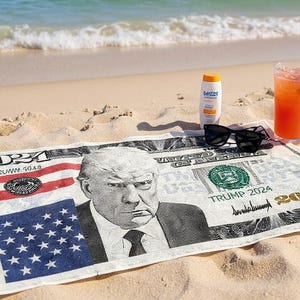 May include: A beach scene with a towel printed to resemble a US bill featuring a portrait. Sunglasses, sunscreen, and a cocktail with a straw are on the sand. The towel has the text "TRUMP 2024".