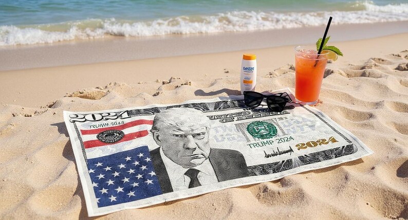 May include: A beach scene with a towel printed to resemble a US bill featuring a portrait. Sunglasses, sunscreen, and a cocktail with a straw are on the sand. The towel has the text "TRUMP 2024".