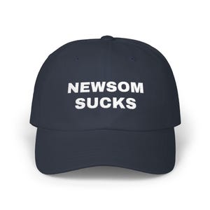 May include: Navy blue baseball cap with the white text "NEWSOM SUCKS" embroidered on the front. The cap has a curved brim and a structured crown. The hat is a classic, casual accessory.