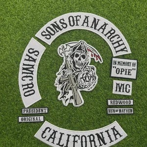 Op de afbeelding: Een verzameling geborduurde patches met de tekst "SONS OF ANARCHY" in een gebogen ontwerp. Andere patches bevatten "SAMCRO", "CALIFORNIA" en een afbeelding van een zeisdrager met een geweer. Extra patches luiden "IN MEMORY OF OPIE", "MC", "REDWOOD", "MEN OF MAYHEM", "PRESIDENT" en "ORIGINAL".