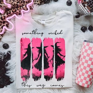 Può includere: T-shirt bianco con effetto sfumato rosa e la scritta "something wicked this way comes". La maglietta presenta una grafica di quattro sagome di streghe nere su pennellate rosa. È visibile un bicchiere a scacchi rosa e bianco.