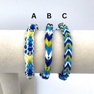 May include: Three rubber band bracelets labeled A, B, and C. Bracelet A features a complex woven pattern in blue, white, and yellow. Bracelet B has a braided design in blue, white, and yellow. Bracelet C displays a thick fishtail weave in blue, white, and yellow.