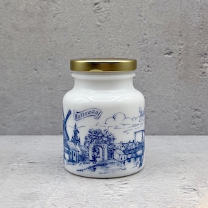 May include: A white ceramic jar with a gold lid, featuring a blue and white illustration of a Dutch village scene. The jar has the word "Wytemaal" in a banner. The jar is sitting on a gray surface.