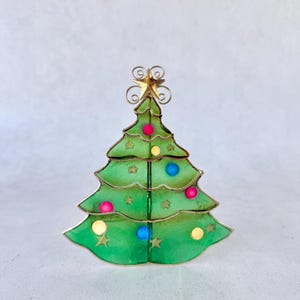 May include: A decorative Christmas tree ornament crafted from green translucent material with a gold wire frame. The tree features small gold star cutouts and round pink, blue, and yellow accents. A gold wire star sits at the very top of the tree.