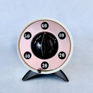 May include: A vintage kitchen timer with a white and pink face, featuring black numbers and a black dial. The timer has a black base and a retro design, perfect for cooking or baking.